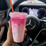 Person holds a pink smoothie cup with an orange straw inside a car, steering wheel and dashboard in the background, pink nails visible on the hand.