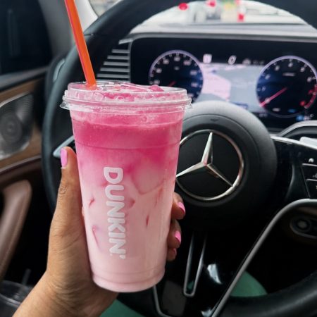 Person holds a pink smoothie cup with an orange straw inside a car, steering wheel and dashboard in the background, pink nails visible on the hand.