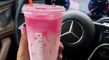 Person holds a pink smoothie cup with an orange straw inside a car, steering wheel and dashboard in the background, pink nails visible on the hand.