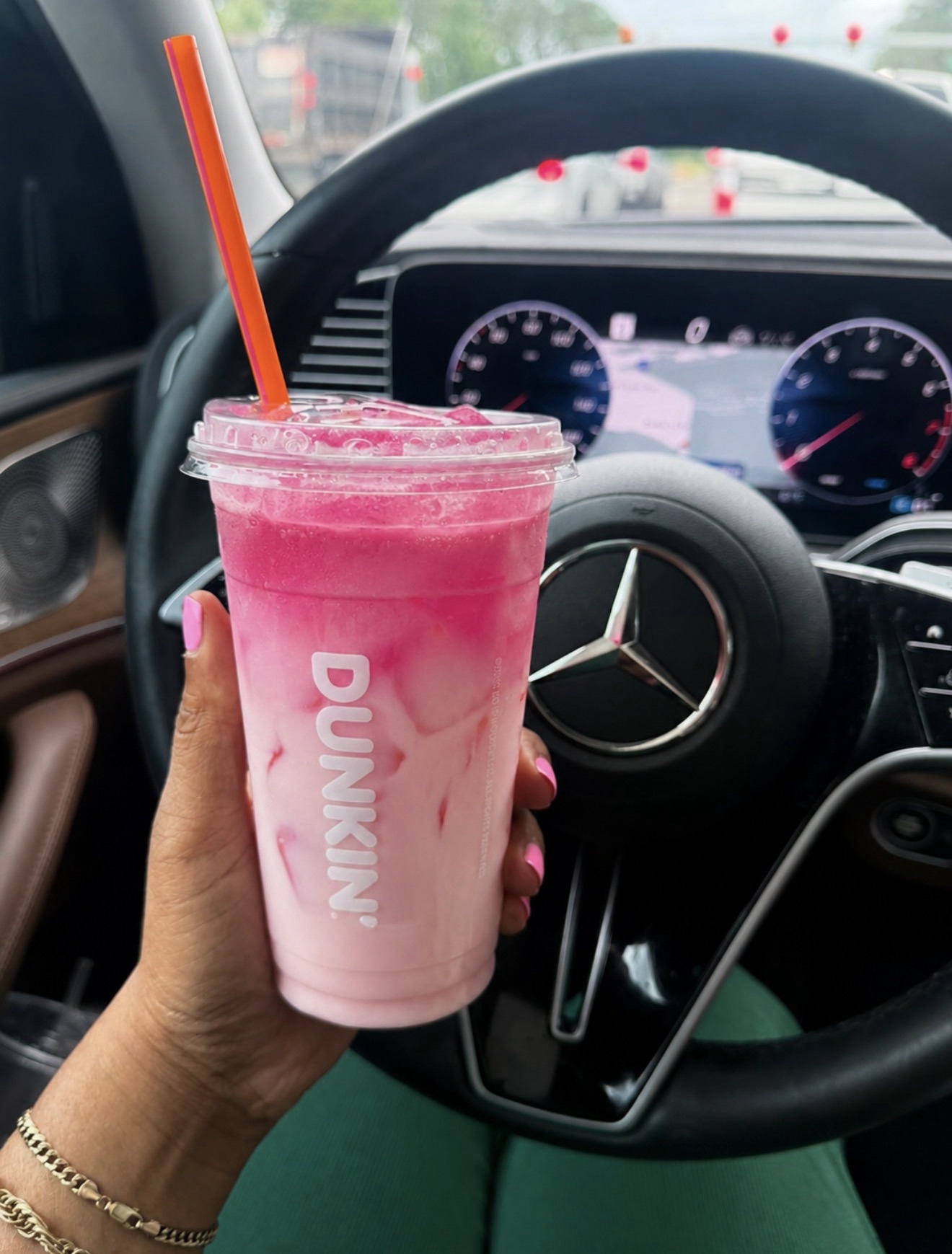 Person holds a pink smoothie cup with an orange straw inside a car, steering wheel and dashboard in the background, pink nails visible on the hand.