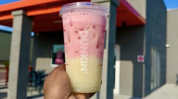 Hand holding a two-toned pink and yellow iced drink in front of a Dunkin' store exterior with a blue sky above.
