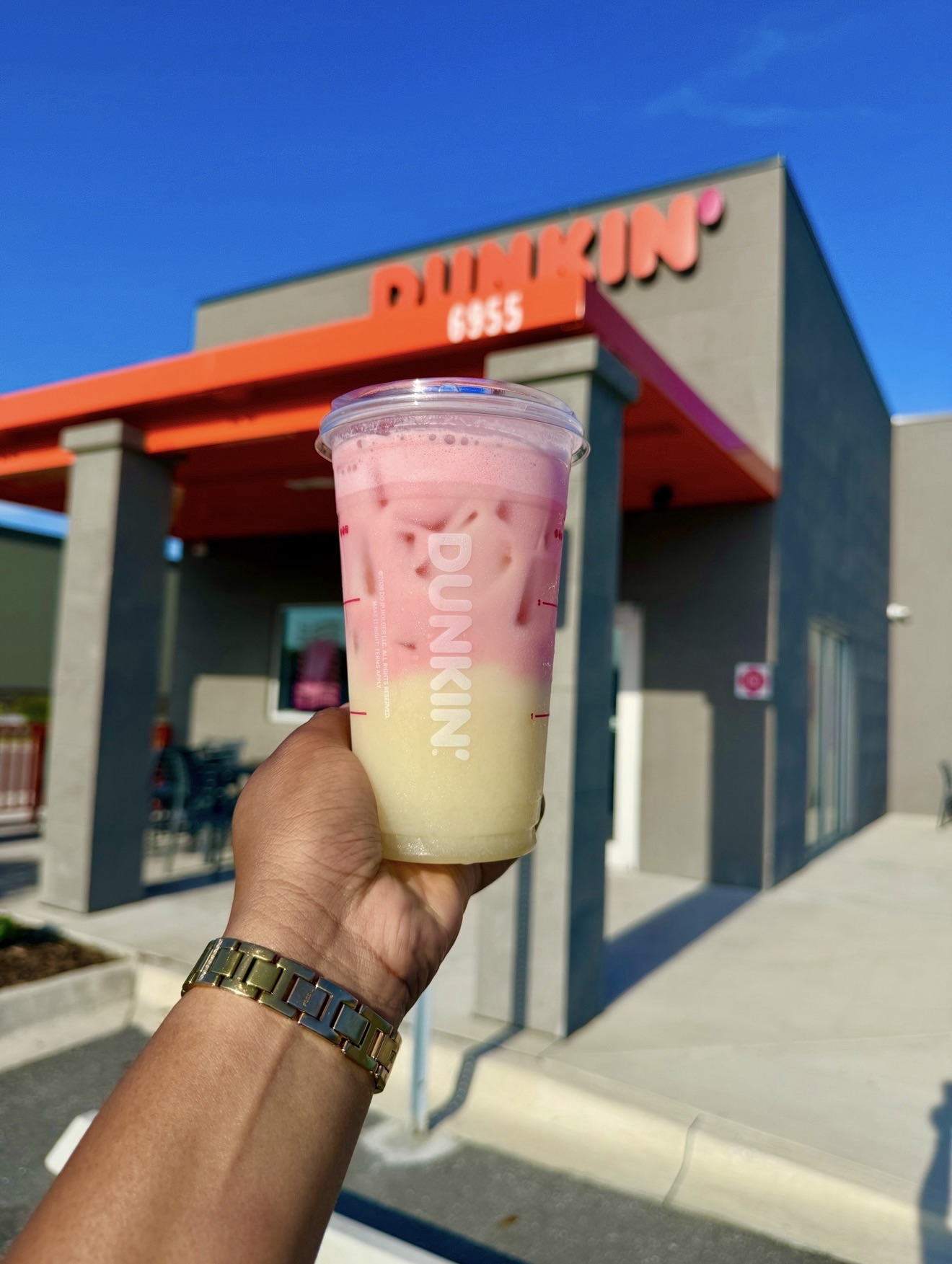 Hand holding a two-toned pink and yellow iced drink in front of a Dunkin' store exterior with a blue sky above.