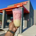 Hand holding a pink-and-cream iced drink in a clear Dunkin' cup in front of a Dunkin' storefront on a sunny day