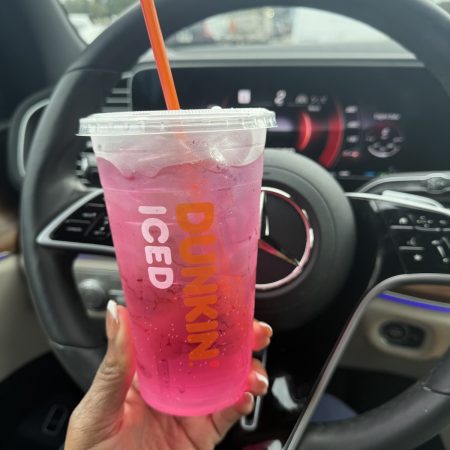 Pink iced Dunkin’ drink in a clear cup with an orange straw, held in front of a car steering wheel and dashboard surface.