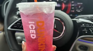 Pink iced Dunkin' drink in a clear cup with an orange straw, held in front of a car steering wheel and dashboard surface.