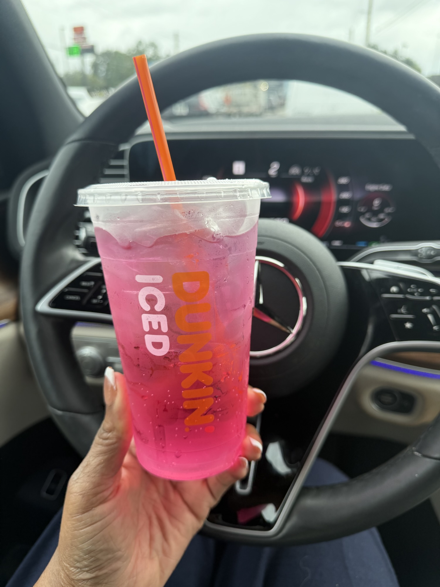 Pink iced Dunkin' drink in a clear cup with an orange straw, held in front of a car steering wheel and dashboard surface.