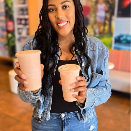 Woman in a denim jacket holding two pink smoothies and smiling at the camera in a cafe setting. 2) Smiling woman presents two pink smoothie cups in a bright cafe. 3) Young woman with wavy hair wearing denim holds pink beverages toward the camera. 4) Two pink smoothie drinks being offered by a woman in a casual cafe. 5) Person in a denim jacket showing two pink beverages in a sunny cafe.