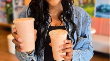 Woman in a denim jacket holding two pink smoothies and smiling at the camera in a cafe setting. 2) Smiling woman presents two pink smoothie cups in a bright cafe. 3) Young woman with wavy hair wearing denim holds pink beverages toward the camera. 4) Two pink smoothie drinks being offered by a woman in a casual cafe. 5) Person in a denim jacket showing two pink beverages in a sunny cafe.