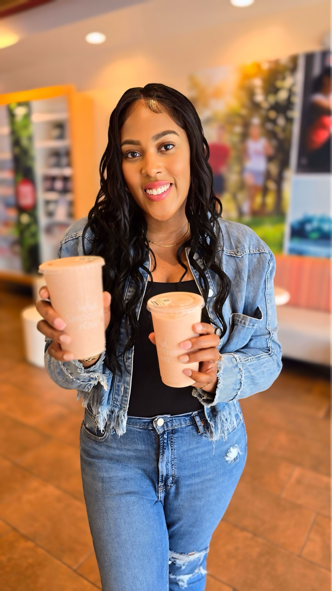 Woman in a denim jacket holding two pink smoothies and smiling at the camera in a cafe setting. 2) Smiling woman presents two pink smoothie cups in a bright cafe. 3) Young woman with wavy hair wearing denim holds pink beverages toward the camera. 4) Two pink smoothie drinks being offered by a woman in a casual cafe. 5) Person in a denim jacket showing two pink beverages in a sunny cafe.