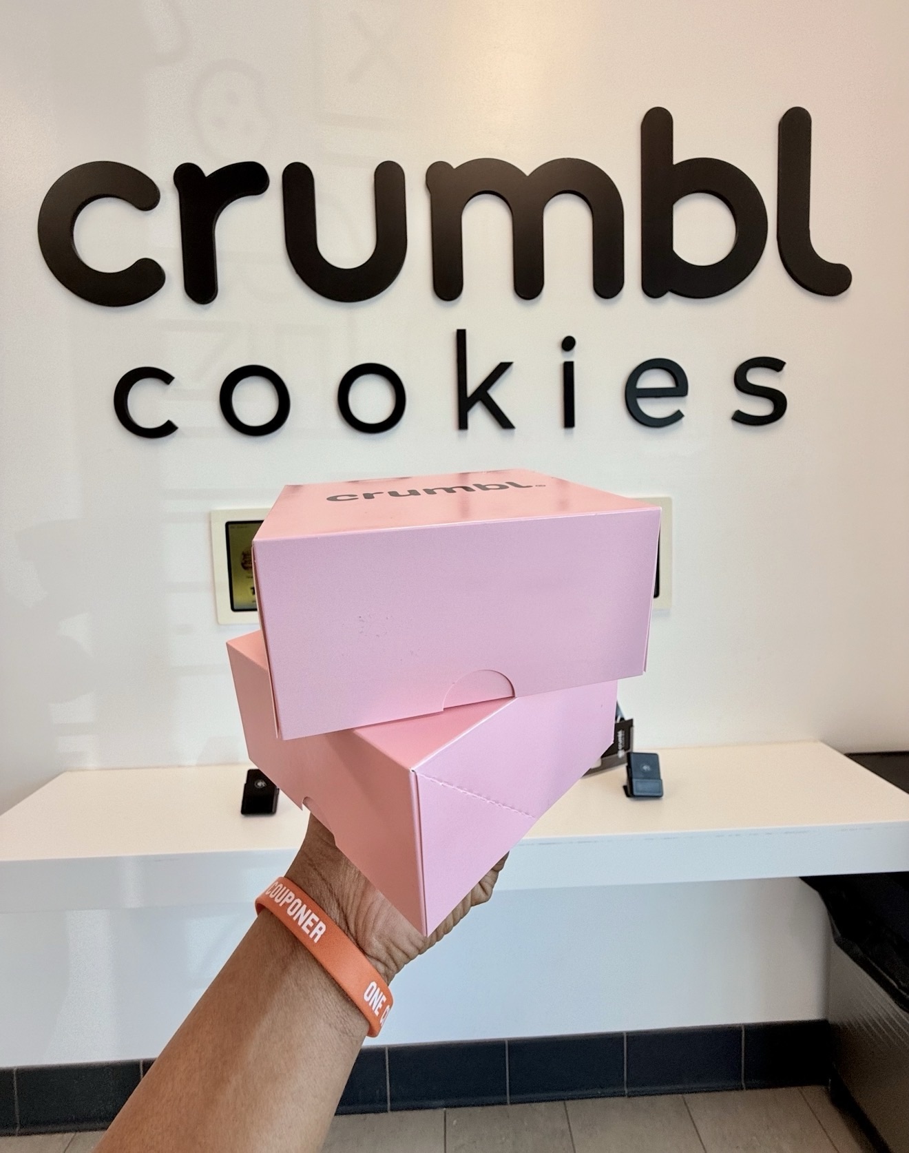 Person holding a pink Crumbl Cookies box in front of a Crumbl Cookies sign on the wall.