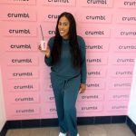 Smiling woman in teal outfit holding a pink Crumbl cup with a straw, standing in front of a pink wall covered with Crumbl logos.