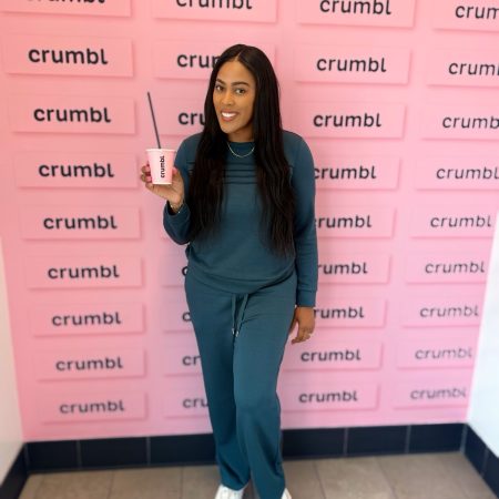 Smiling woman in teal outfit holding a pink Crumbl cup with a straw, standing in front of a pink wall covered with Crumbl logos.