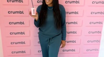 Smiling woman in teal outfit holding a pink Crumbl cup with a straw, standing in front of a pink wall covered with Crumbl logos.