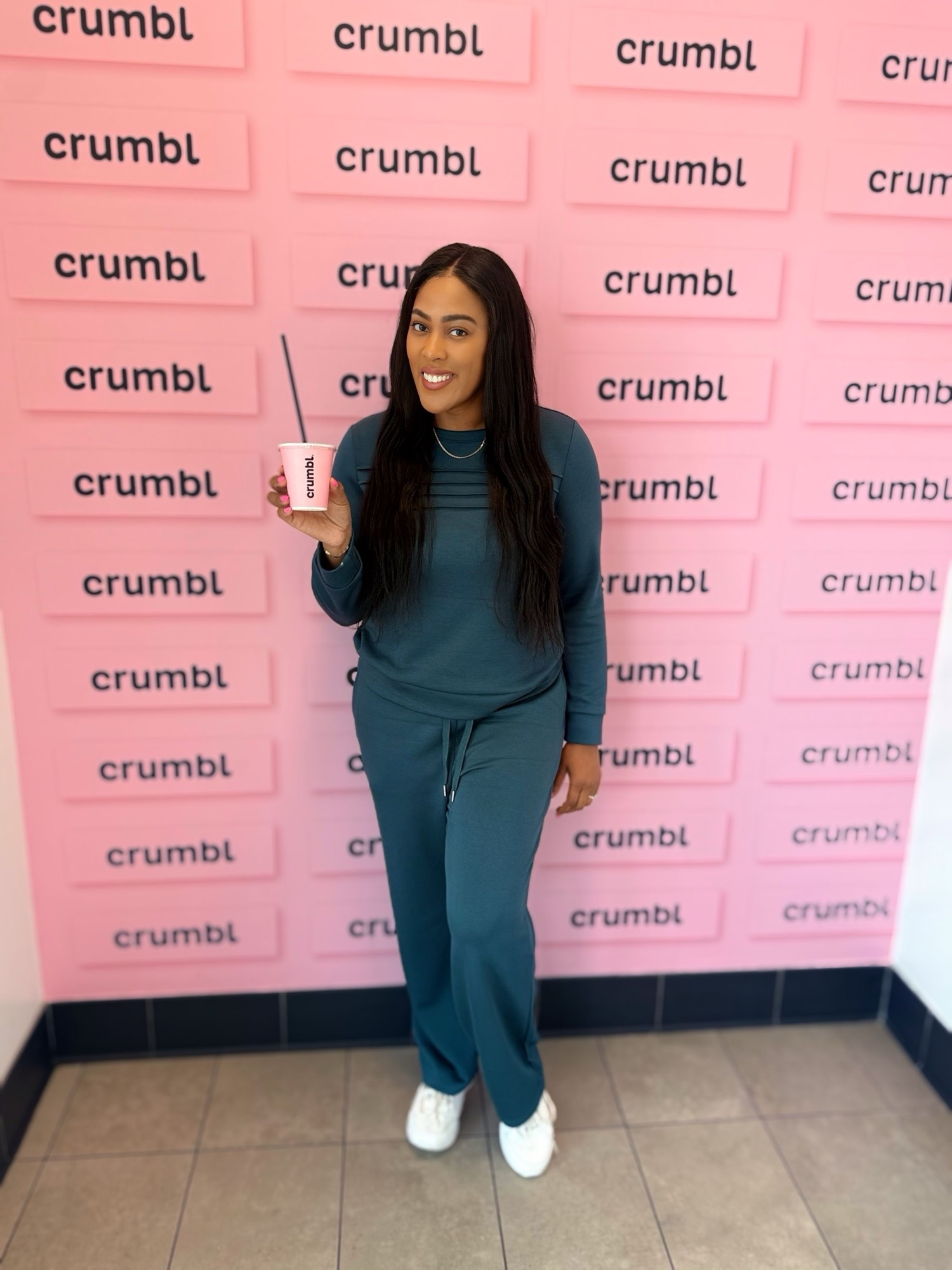 Smiling woman in teal outfit holding a pink Crumbl cup with a straw, standing in front of a pink wall covered with Crumbl logos.